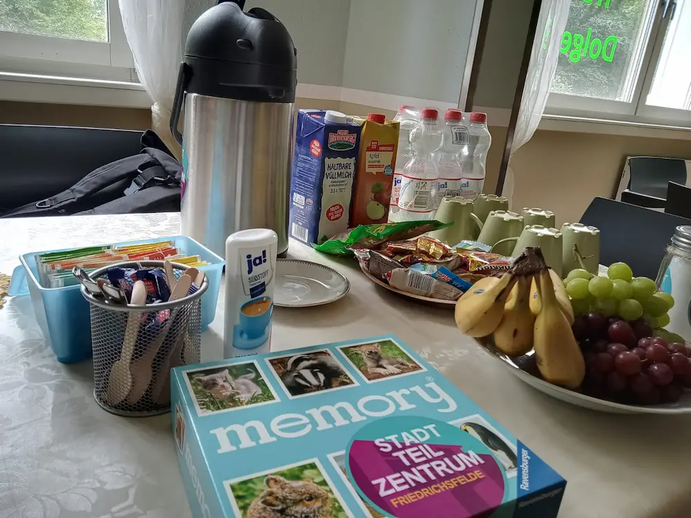 Es sind zu sehen ein Tisch, der mit einer Kaffeekanne, Obst und Snacks gedeckt ist. Davor liegt das Spiel Memory