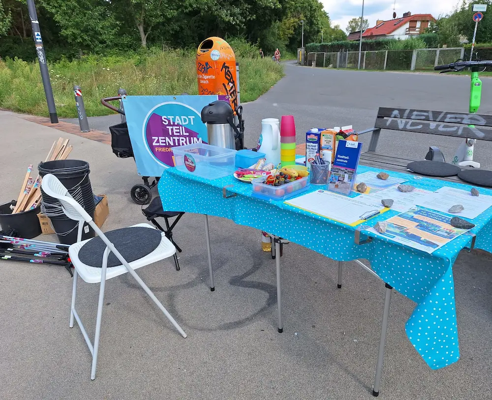 Kiezputz draußen: Tisch mit Infomaterial, Kaffee, Tee, Snacks, dahinter eine Bank und Stühle. Es sind Metalgreifzanger und Eimer zum Müllsammeln zu sehen.