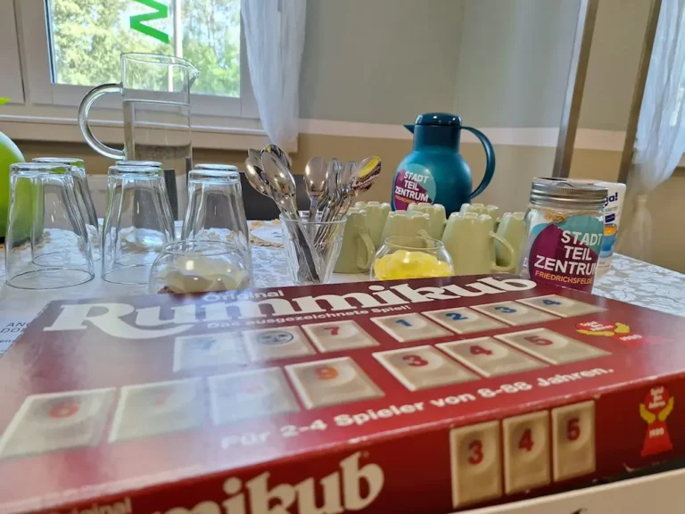 Auf einem Tisch sieht man Gläser und Tassen und eine Kaffeekanne. Davor liegt das Spielrummikub.